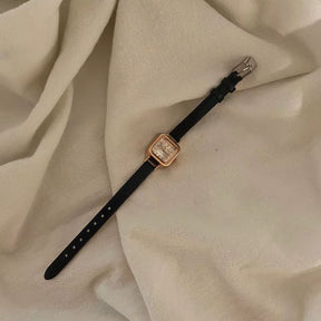 Relógio feminino com pulseira preta de couro e mostrador quadrado dourado.