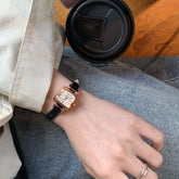 Relógio feminino com pulseira preta de couro e mostrador quadrado dourado.