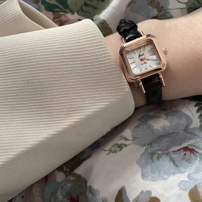 Relógio feminino com pulseira preta de couro e mostrador quadrado dourado.