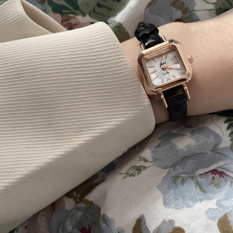 Relógio feminino com pulseira preta de couro e mostrador quadrado dourado.